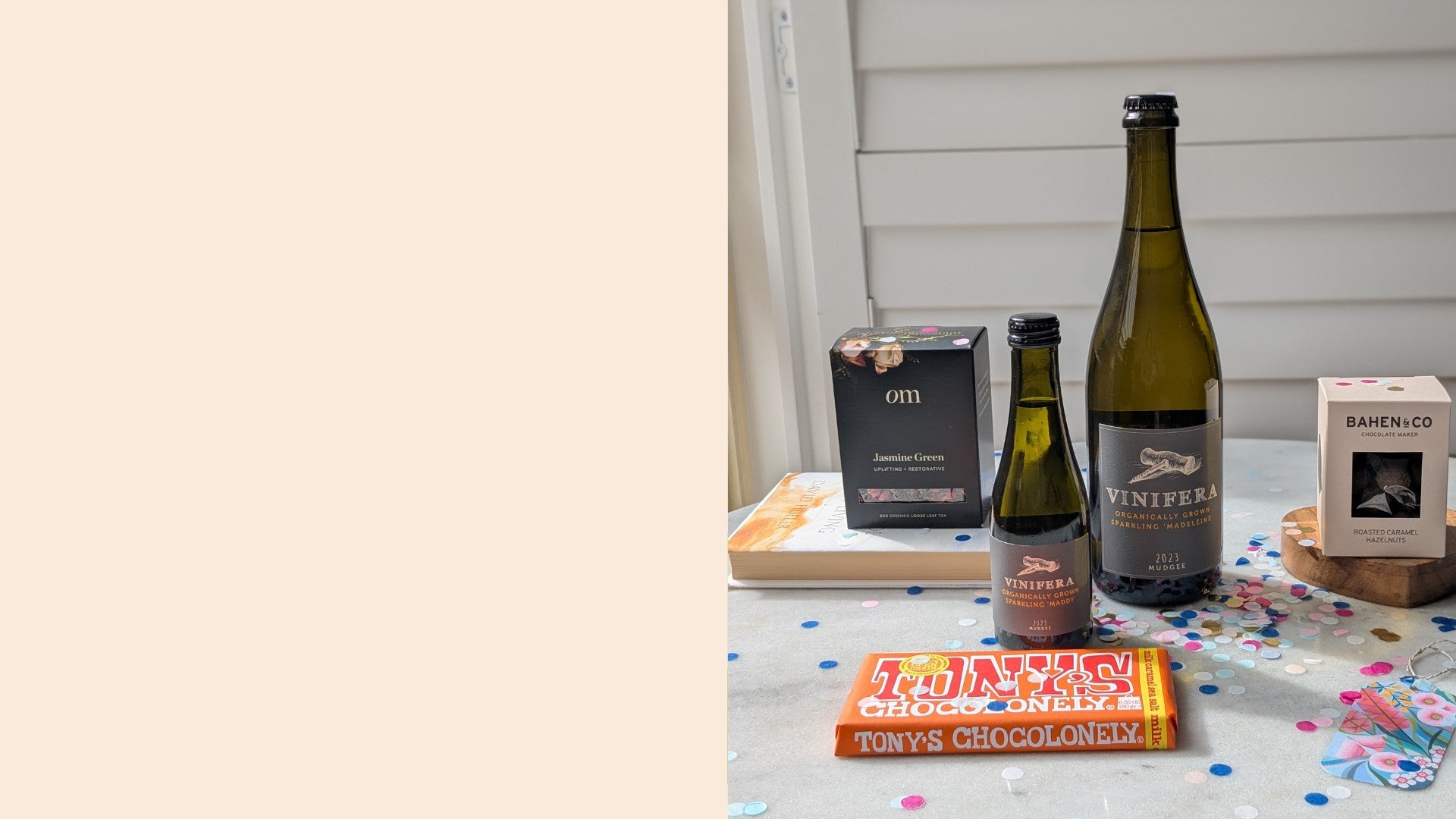 Box of tea leaves, block of chocolate, a small and large bottle of wine, box of coated hazelnuts, scattered confetti, floral gift card on a marble table