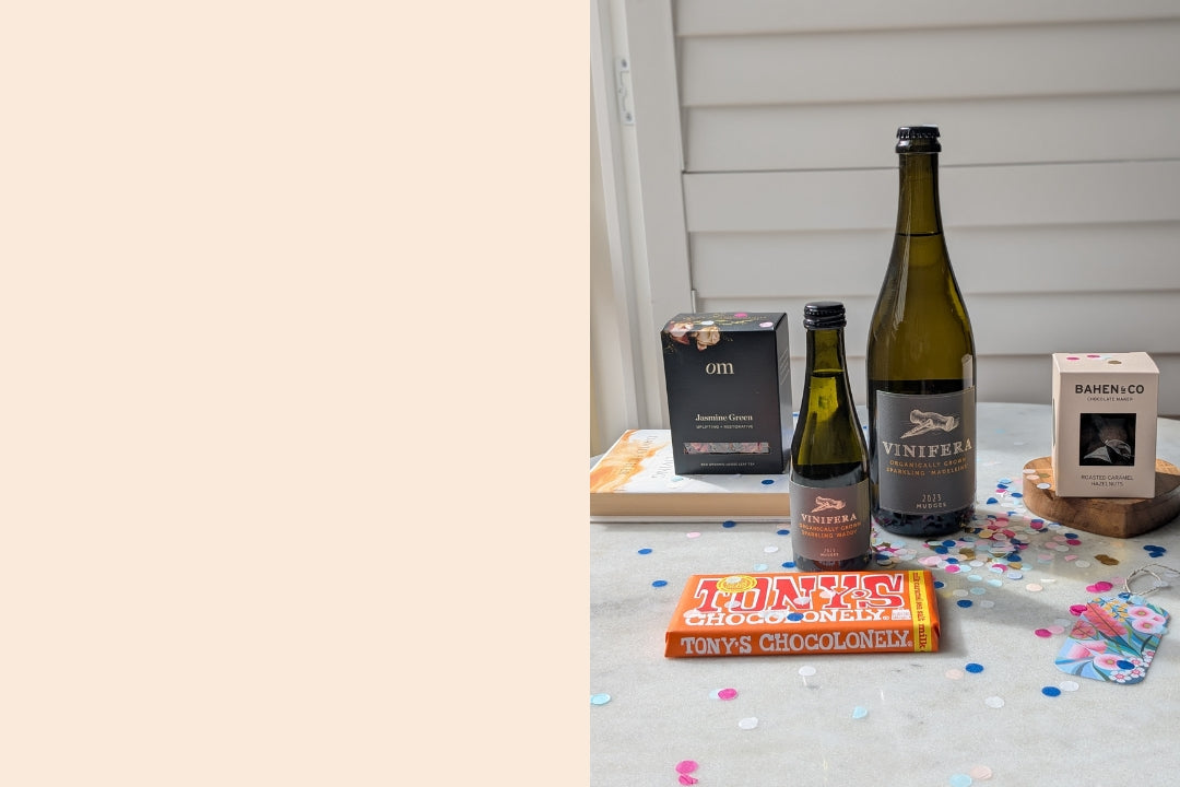 Box of tea leaves, block of chocolate, a small and large bottle of wine, box of coated hazelnuts, scattered confetti, floral gift card on a marble table